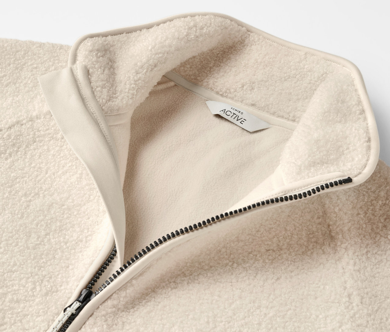 Egy bézs színű, cipzáros, galléros fleece pulóver részlete.