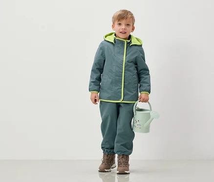 Egy fiú kabátban és nadrágban világoszöld locsolókannát tart.