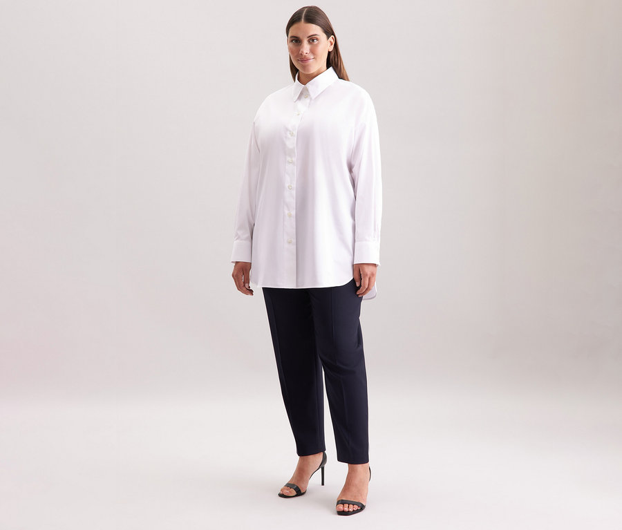 Nő visel egy fehér Seidensticker túlméretes hosszú ingblúzt, sötét nadrágot és fekete szandálokat.