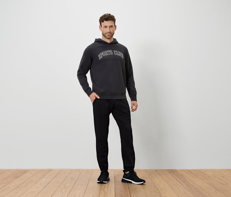 Férfi áll a fapadlón, sötétszürke kapucnis pulóvert és fekete nadrágot visel.