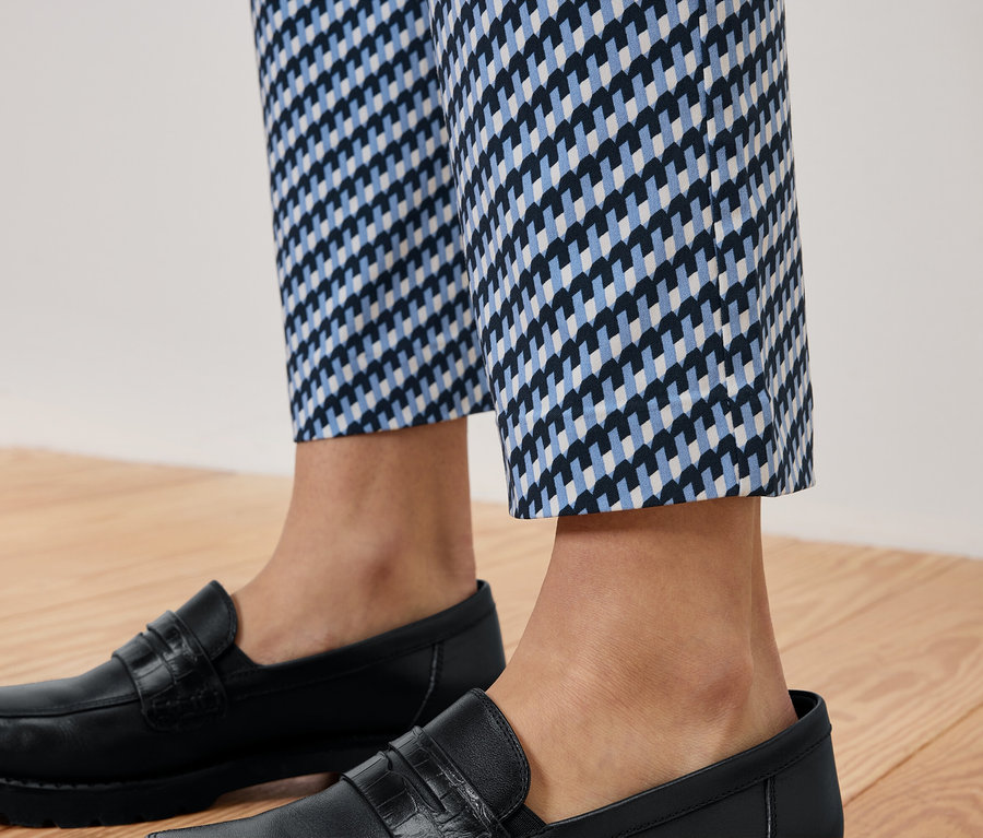 Egy személy lábai kék-fehér mintás nadrágban és fekete bőr mokaszinban, fa padlón.