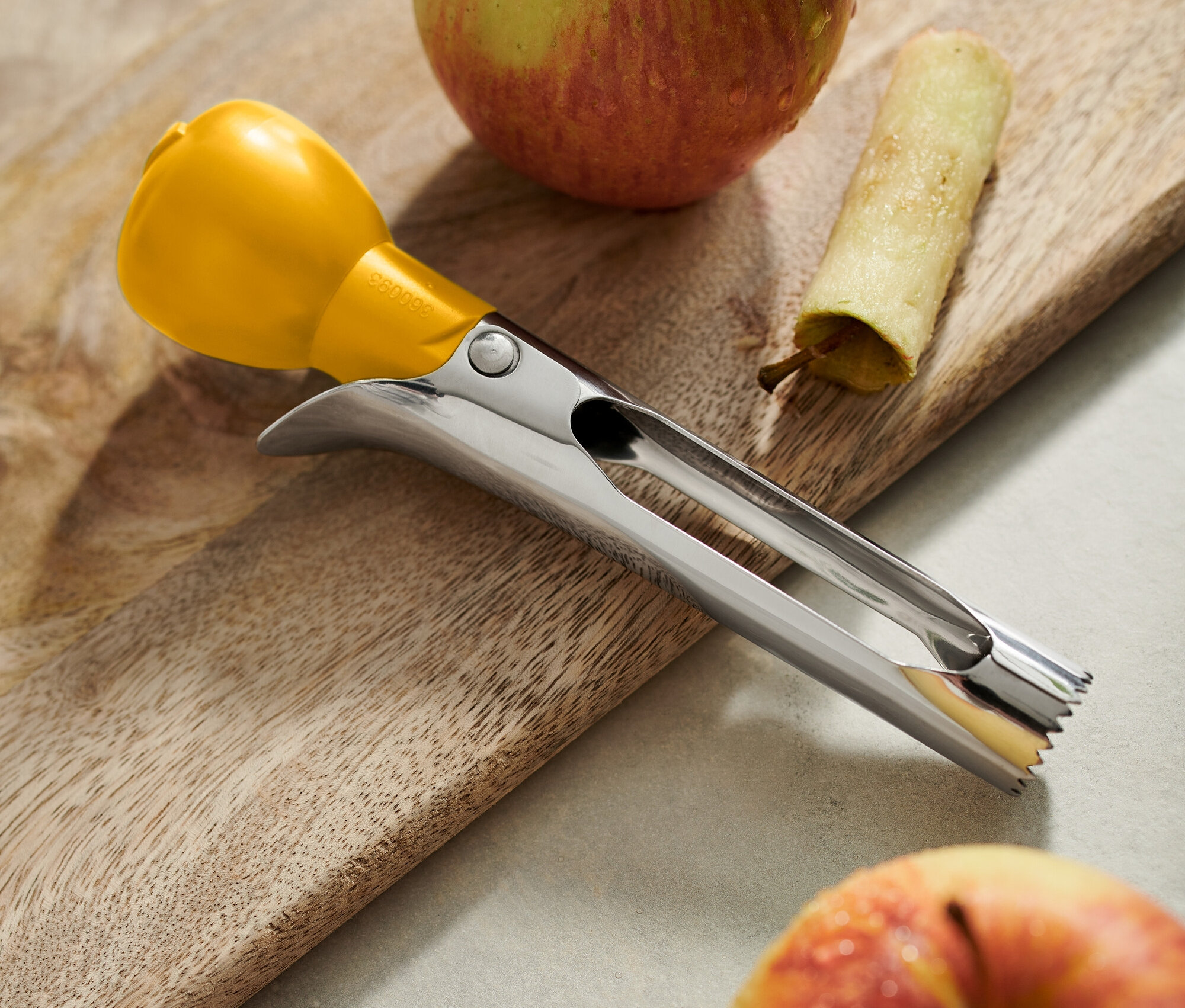 Egy fából készült deszkán két alma fekszik, az egyiknek kivájták a közepét, mellette egy darab héj és egy rozsdamentes acél magozó sárga fogantyúval.
