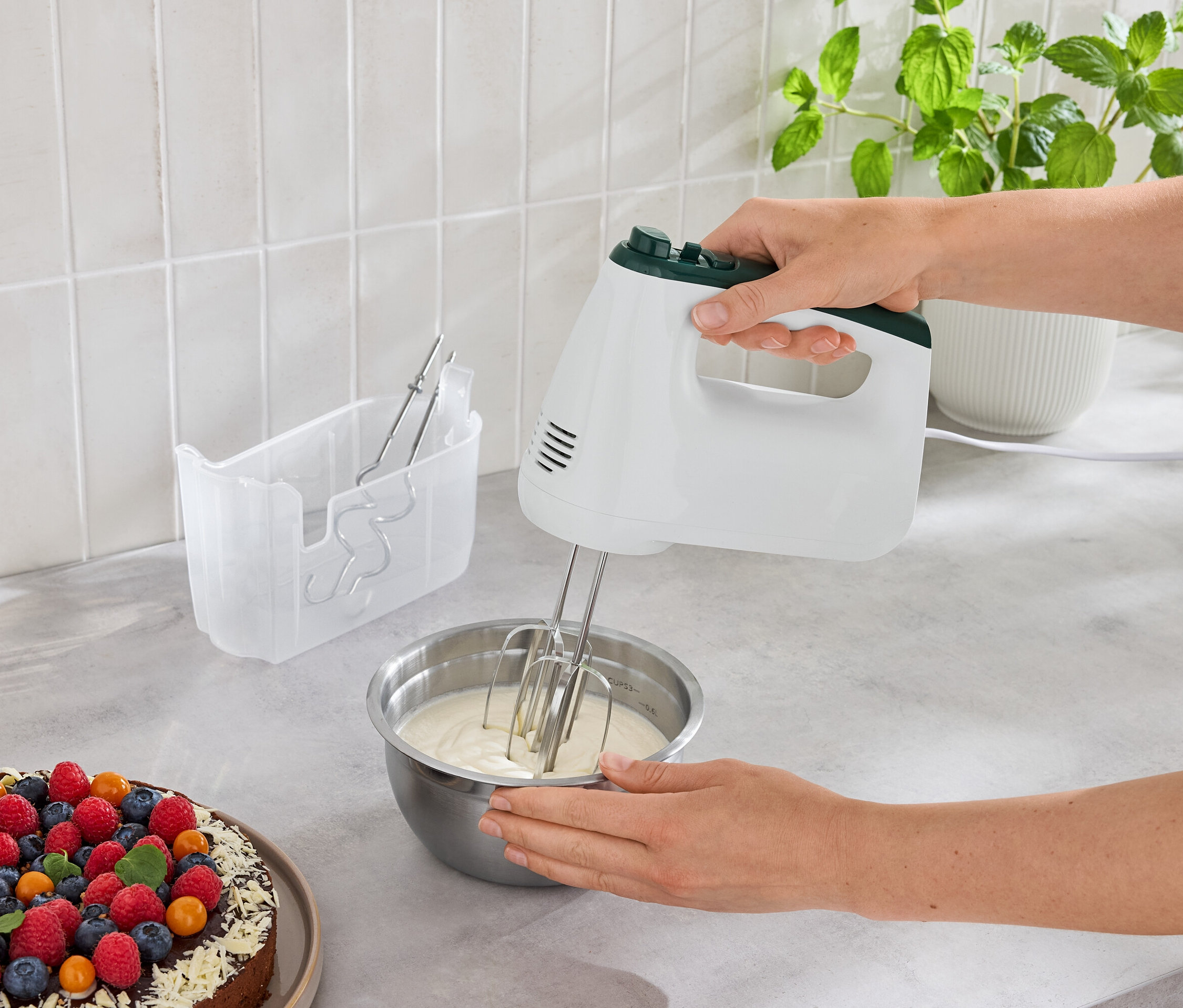 Egy nő tésztát kever egy tálban egy kézi mixerrel egy gyümölcsös torta mellett.