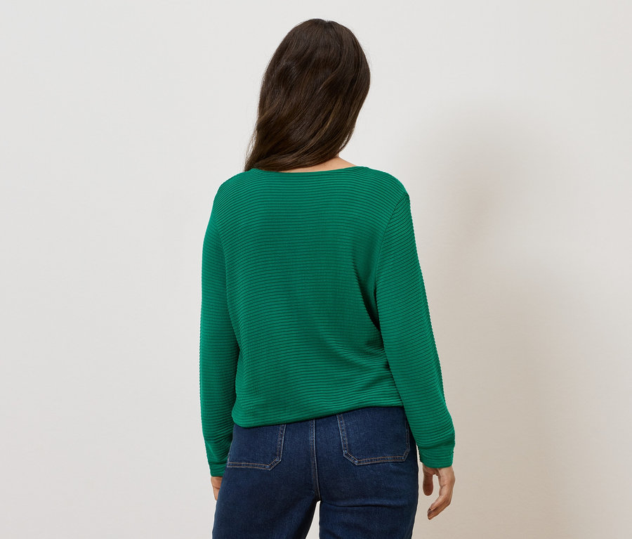 Egy nő áll háttal, zöld pulóvert és farmert visel.