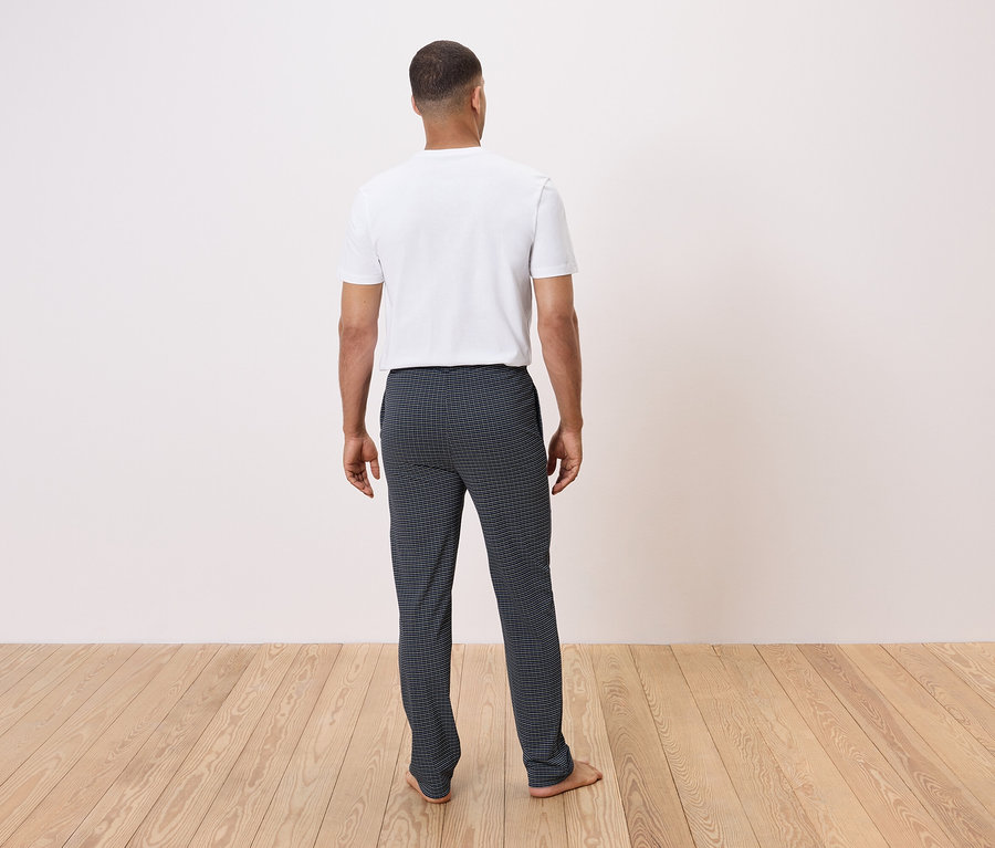 Egy férfi háttal a kamerának áll egy fa padlón, fehér pólót és sötét kockás nadrágot visel.