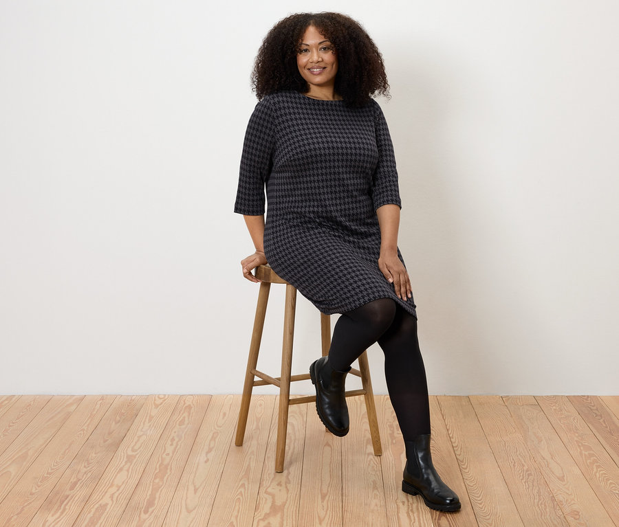 Nő ül egy széken, és jacquard tyúklábmintás jerseyruhát visel.