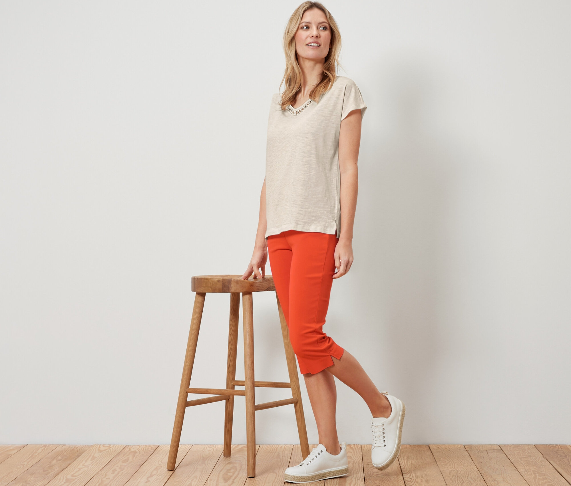 Szőke nő pózol egy fa szék mellett narancssárga 3/4-es sztreccsnadrágban.