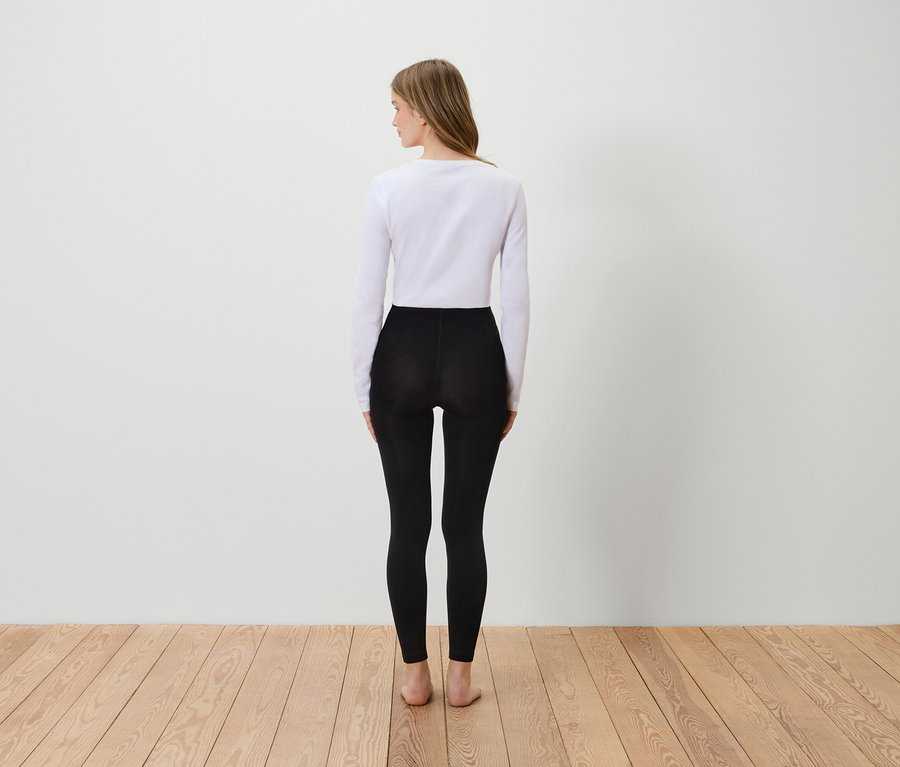 Egy nő hátulról áll, fehér hosszú ujjú inget és fekete leggingset visel.