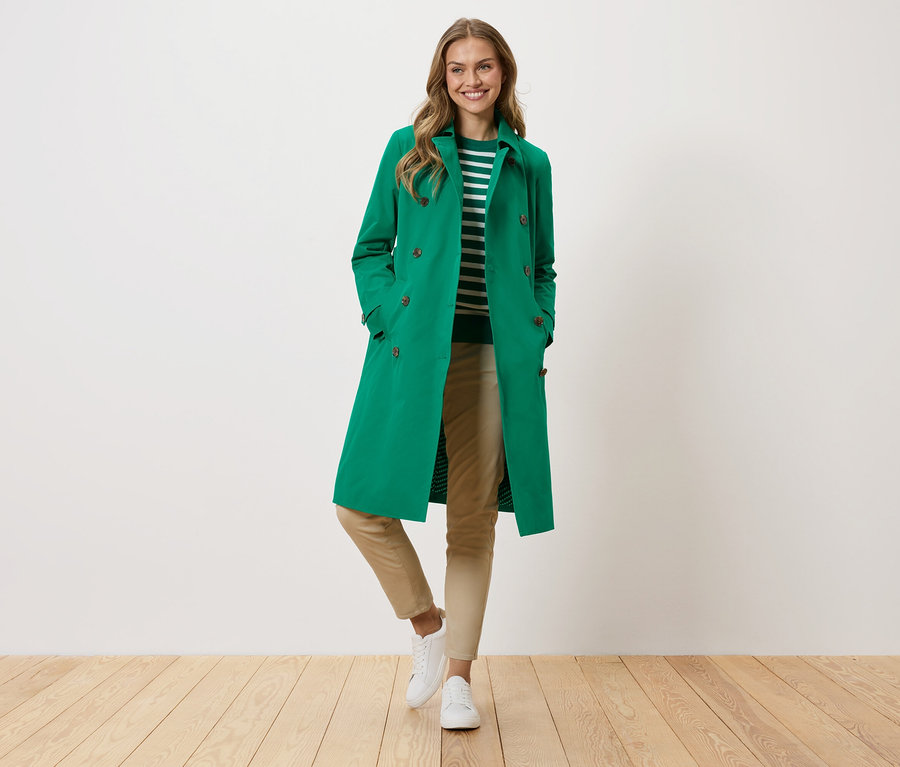 Nő zöld kabátban, csíkos pulóverben, bézs nadrágban és fehér tornacipőben áll egy fehér fal előtt.