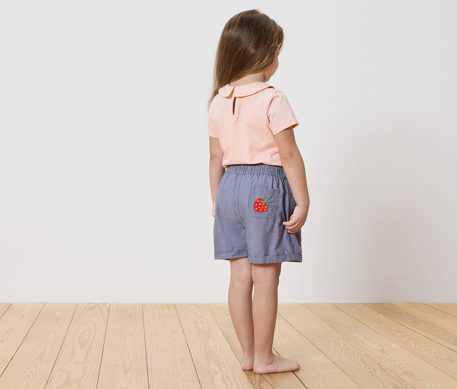 Hátulról látható lány rózsaszín ingben és kék, epraj alkalmazásos rövidnadrágban.