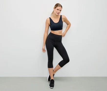 Nő fekete sportmelltartóban és fekete leggingsben pózol egy fehér fal előtt.