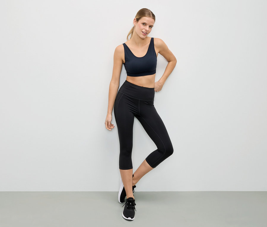 Nő fekete sportmelltartóban és fekete leggingsben pózol egy fehér fal előtt.