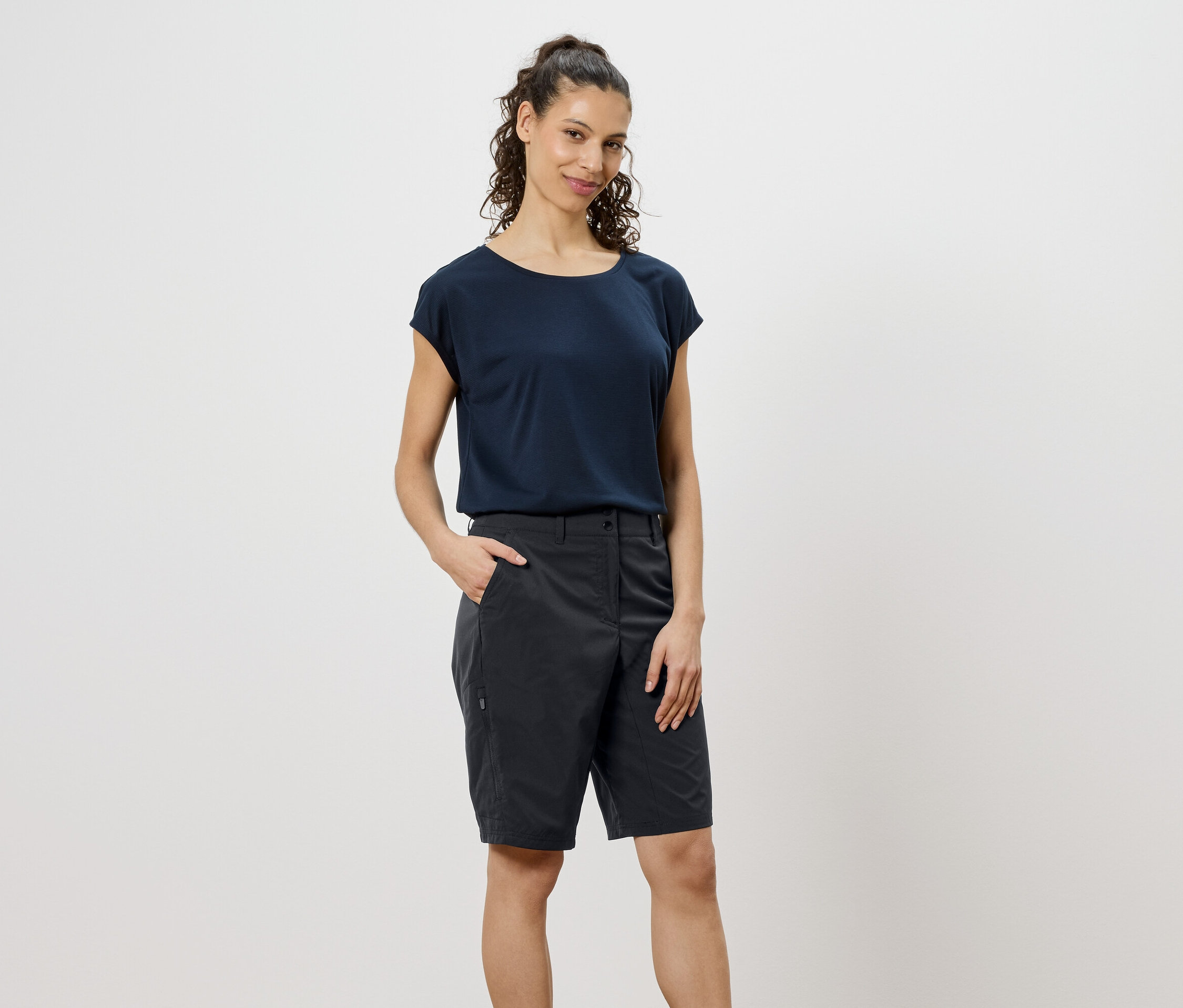 Göndör, barna hajú nő sötétkék pólót és szürke rövidnadrágot visel, kezét a zsebébe dugva.