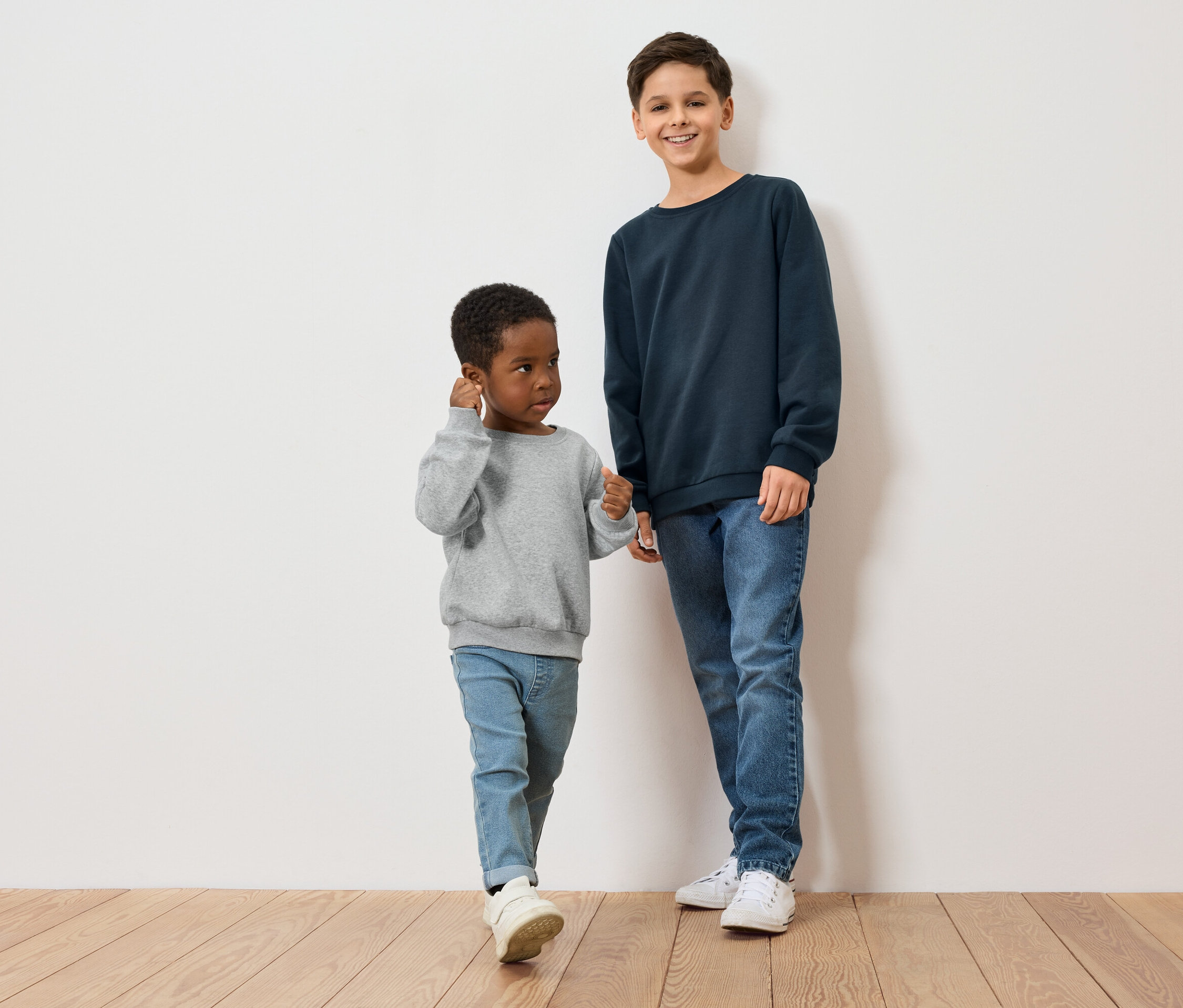 Két fiú, egy sötét bőrű szürke pulóverben, a másik világos bőrű kék pulóverben áll egy fapadlón egy fehér fal előtt.