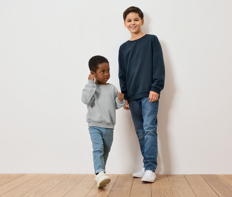 Két fiú, egy sötét bőrű szürke pulóverben, a másik világos bőrű kék pulóverben áll egy fapadlón egy fehér fal előtt.