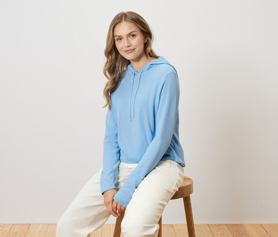 Egy hosszú szőke hajú nő ül egy fa széken, világoskék kapucnis pulóvert és fehér nadrágot visel.