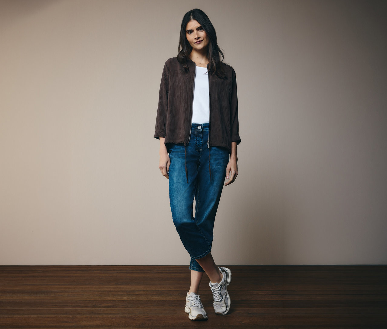 Nő fehér pólóban, farmerben és barna CECIL bluzonban. Egy fal előtt áll.