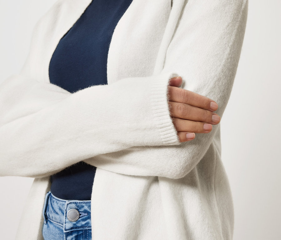 A karjait keresztbe tevő nő hosszú, fehér kardigánt visel egy sötétkék felső és kék farmer felett.