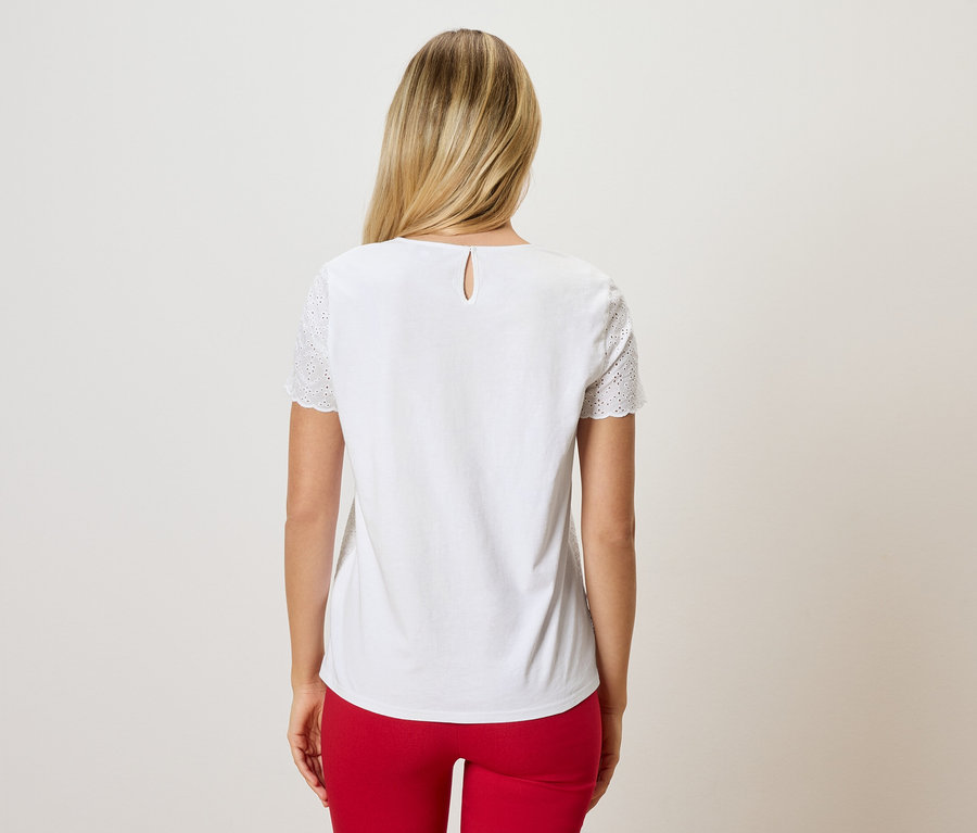 Hátulnézet egy szőke hajú nőről, aki fehér, hímzett ujjú blúzt és piros nadrágot visel.