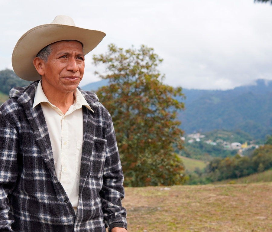 Egy idős férfi világos cowboykalapban és fekete-fehér kockás kabátban áll a szabadban, jobbra nézve. Mögötte zöld hegyek, egy völgy faluval és borús égbolt látható.