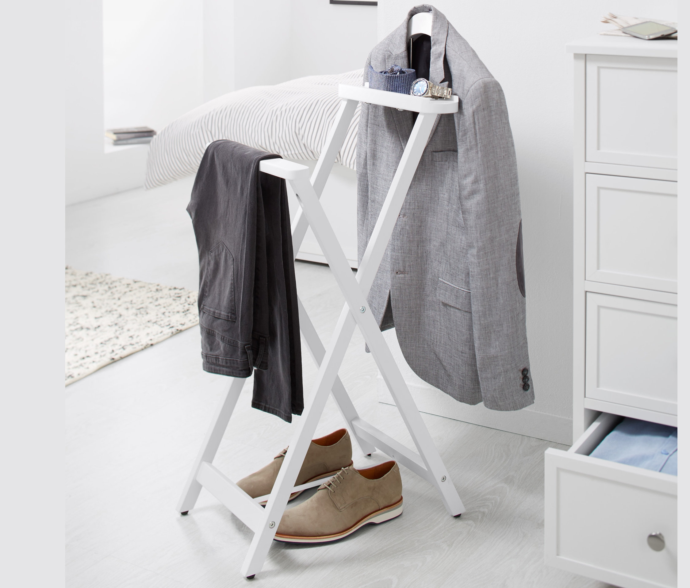 Fehér háttér előtt egy összecsukható, fehér szobainas látható, rajta egy szürke blézer és egy nadrág lóg, alatta barna cipők állnak.