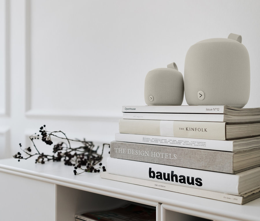 Két különböző méretű szürke Bluetooth® hangszóró egymáson fekszik egy könyvkupacon egy fehér polcon.