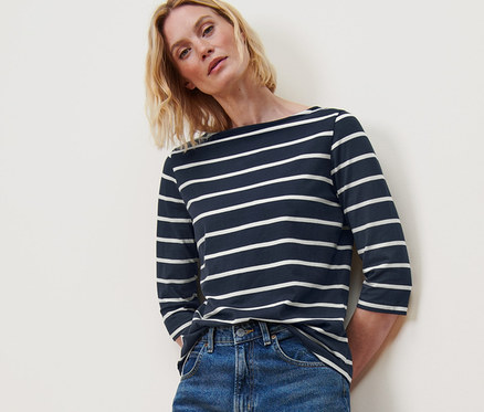 Nő pózol egy 3/4-es ujjú, csíkos felsőben, kék farmerben és fehér tornacipőben.