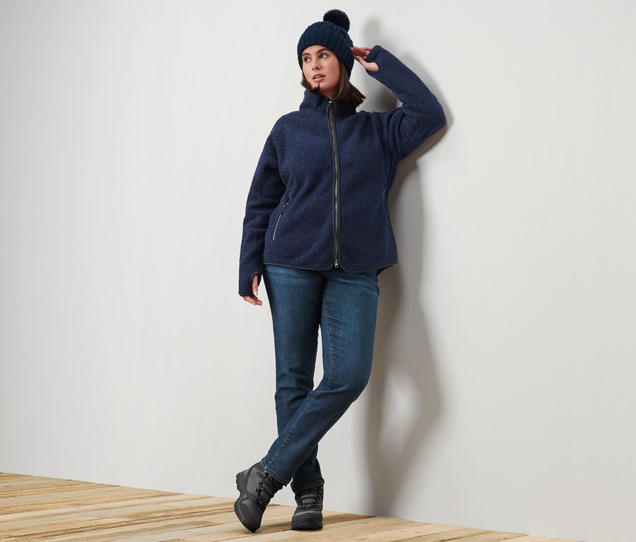 Egy nő a falnak dőlve pózol, kék plüsskabátot, sötétkék Slim Fit Emma farmert és kötött sapkát visel.