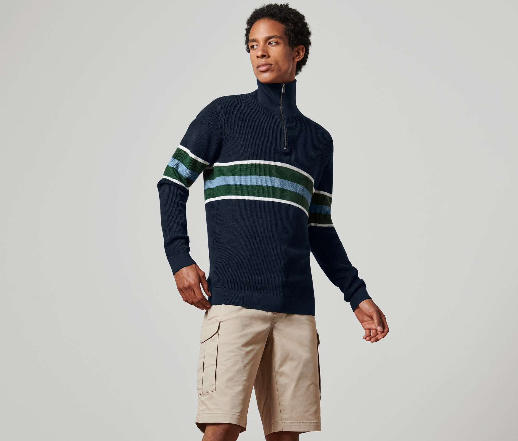 Férfi sötétkék, csíkos, állógalléros férfi pulóvert és khaki rövidnadrágot visel.