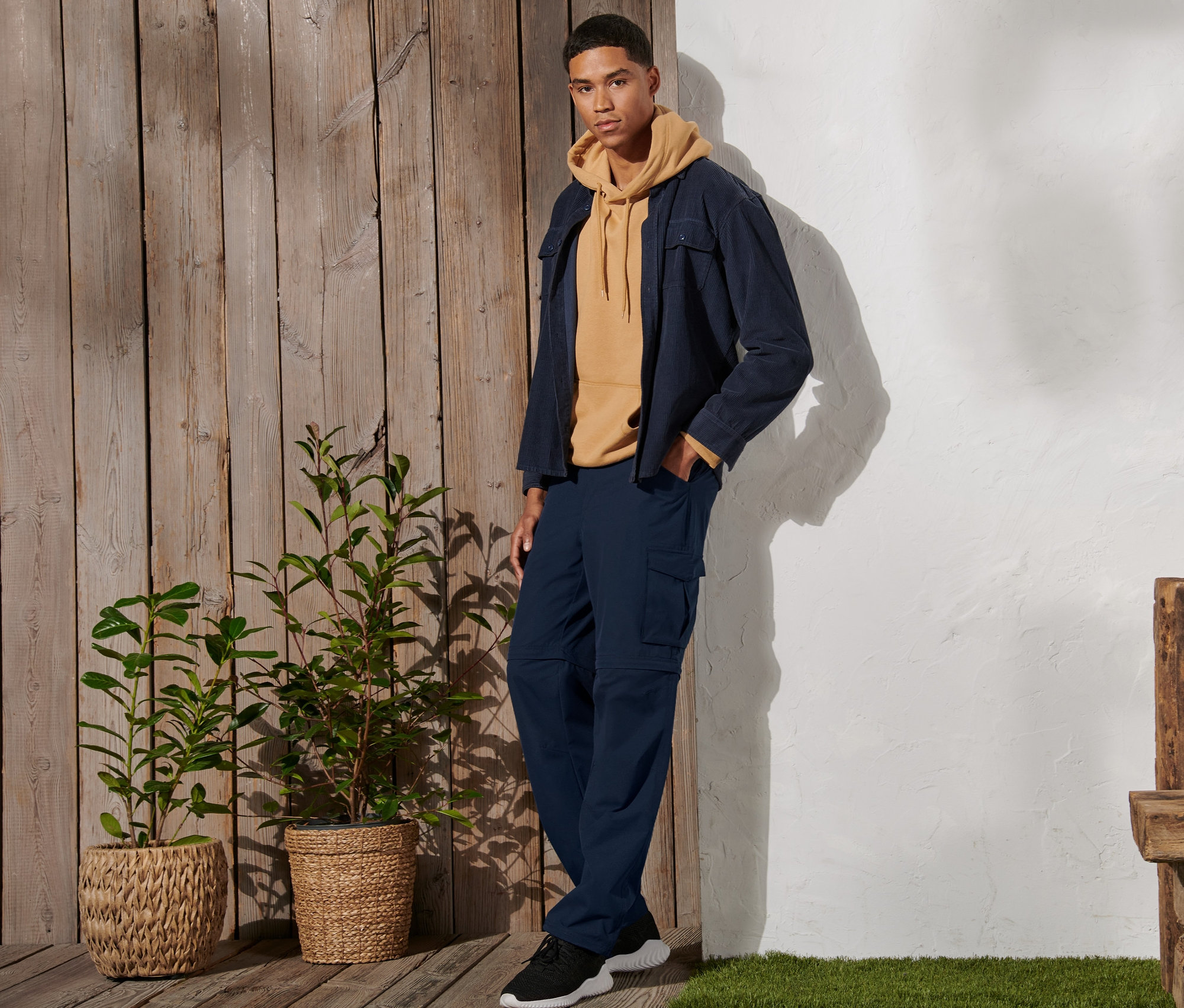 Férfi áll és sötétkék 2 az 1-ben funkcionális nadrágot, sárga kapucnis pulóvert és kabátot visel.