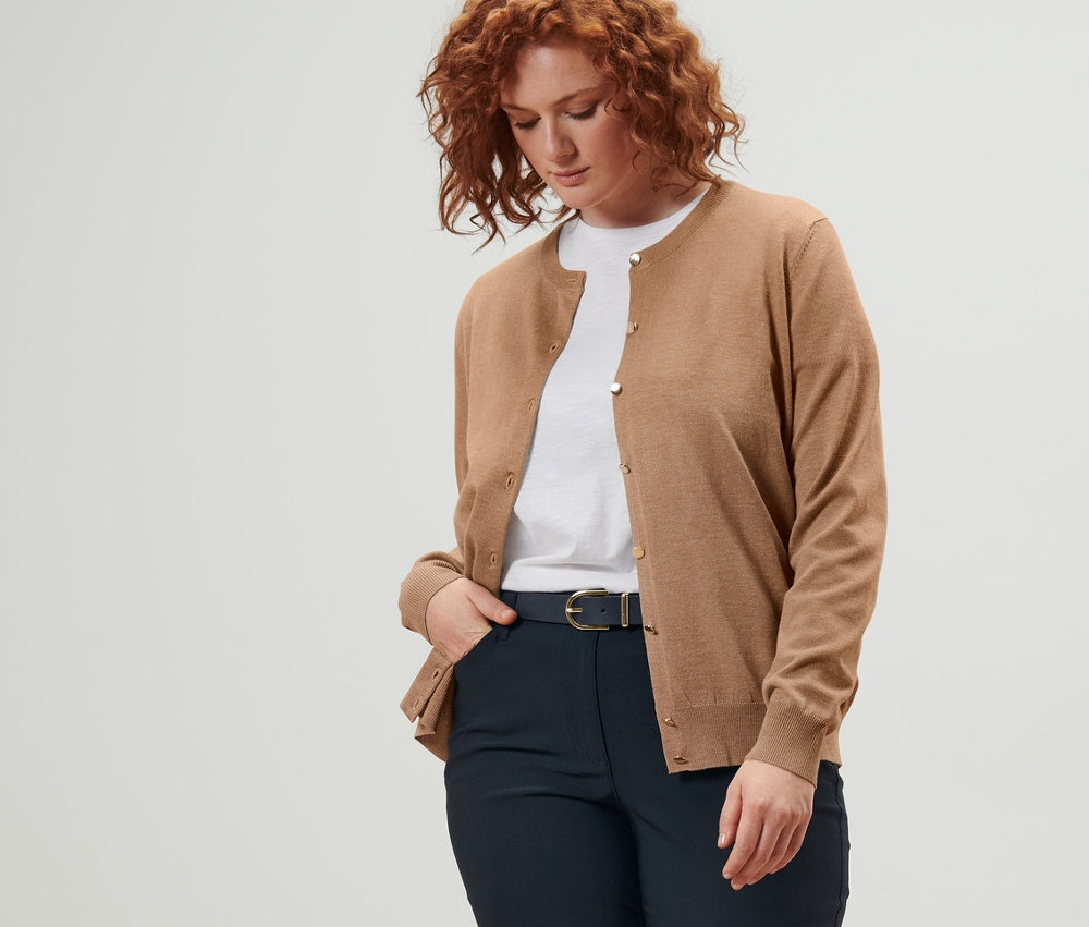 Nő visel bézs finomkötésű kardigánt, sötétkék bőrövet és sötétkék bengalin nadrágot.