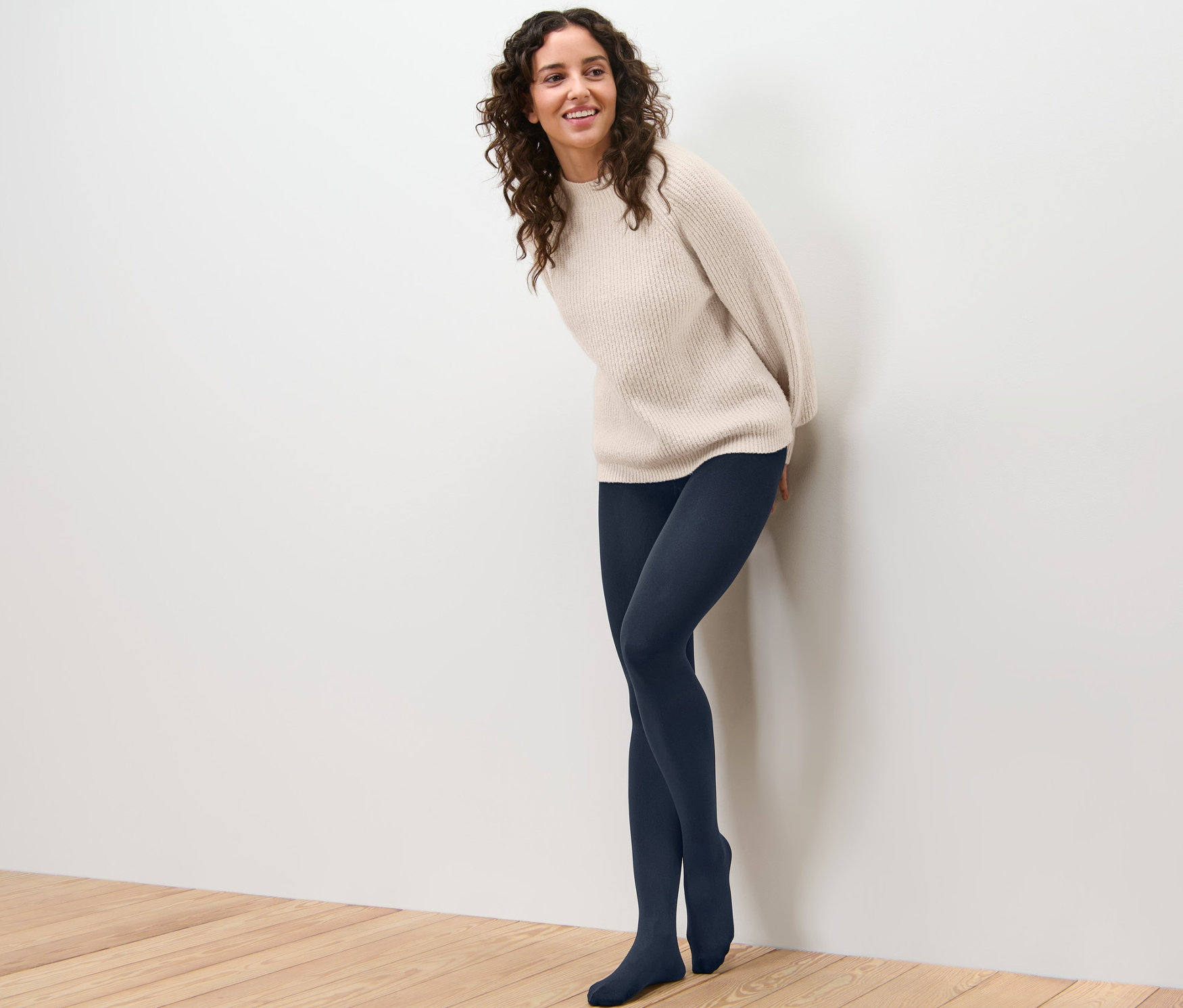Mosolygó nő a falnak dőlve sötétkék termo harisnyát és pulóvert visel.