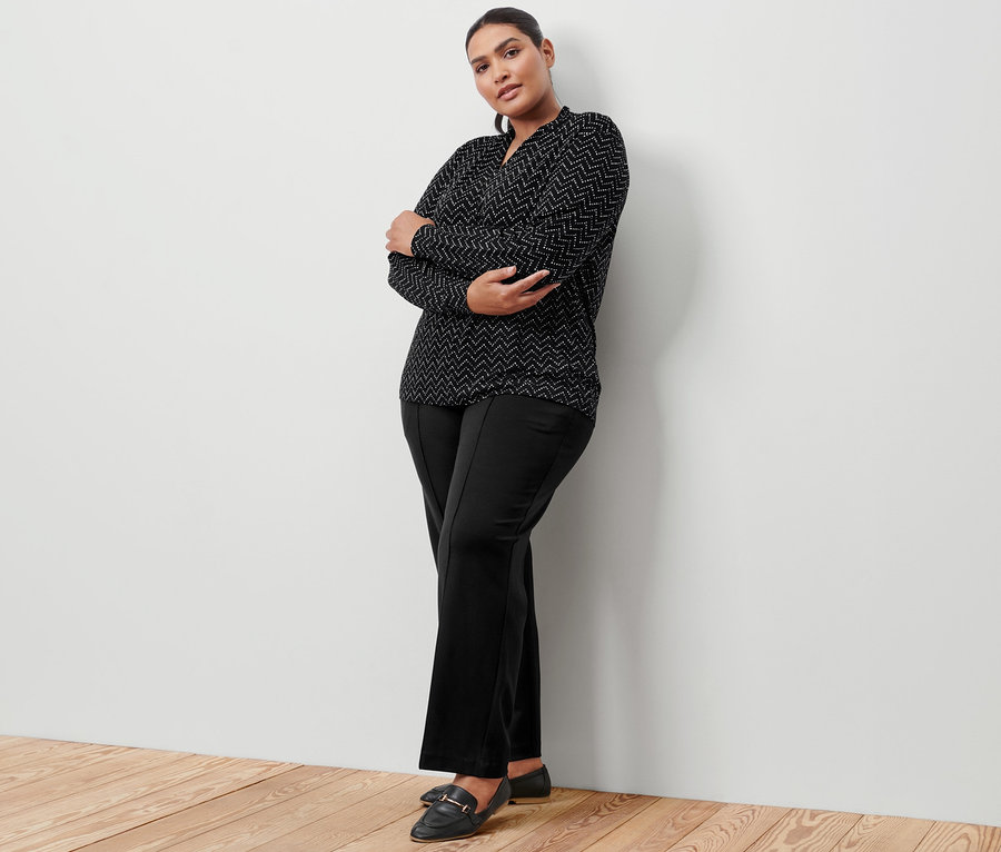 Nő pózol fekete dzsörzénadrágban levarrt hajtással és nyomtatott mintás, V-nyakkivágású hosszú ujjú felsőben.
