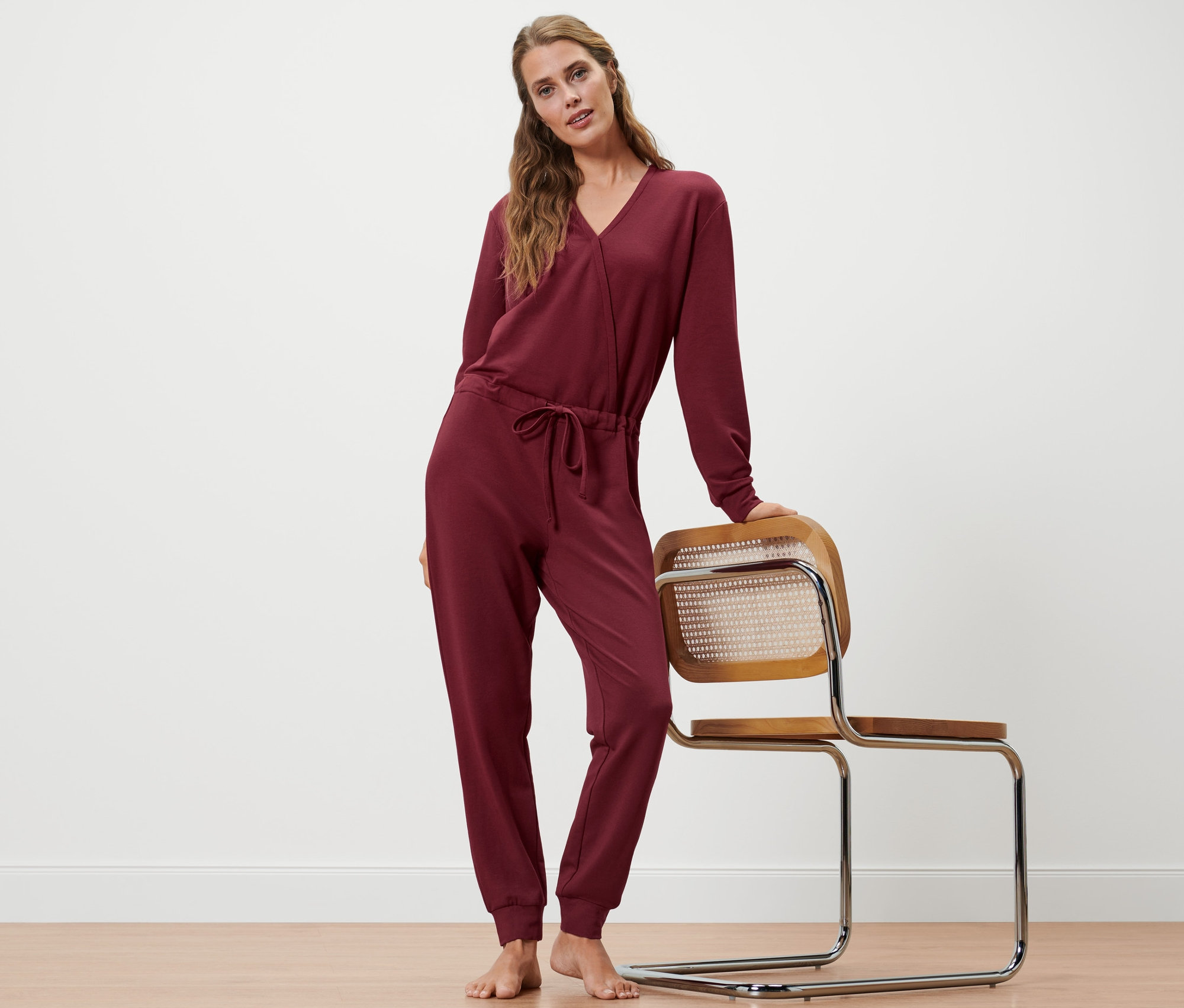 Nő pózol bordó színű női kezeslábasban, egy széken pihentetve a kezét.