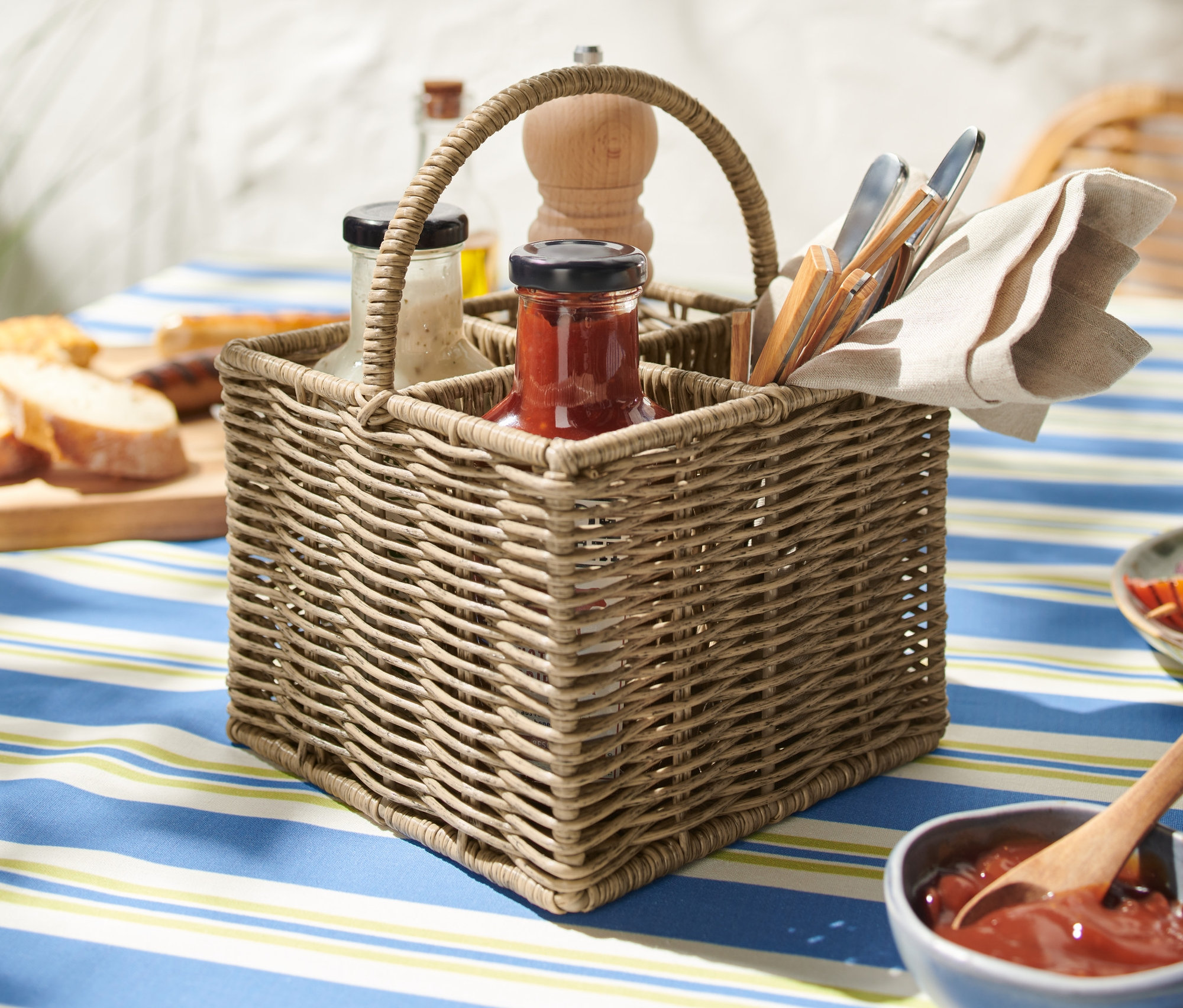 Egy fonott evőeszköztartó kosár kék-fehér csíkos terítőn. A kosárban poharak, borsőrlő, szalvéta és evőeszközök találhatók.