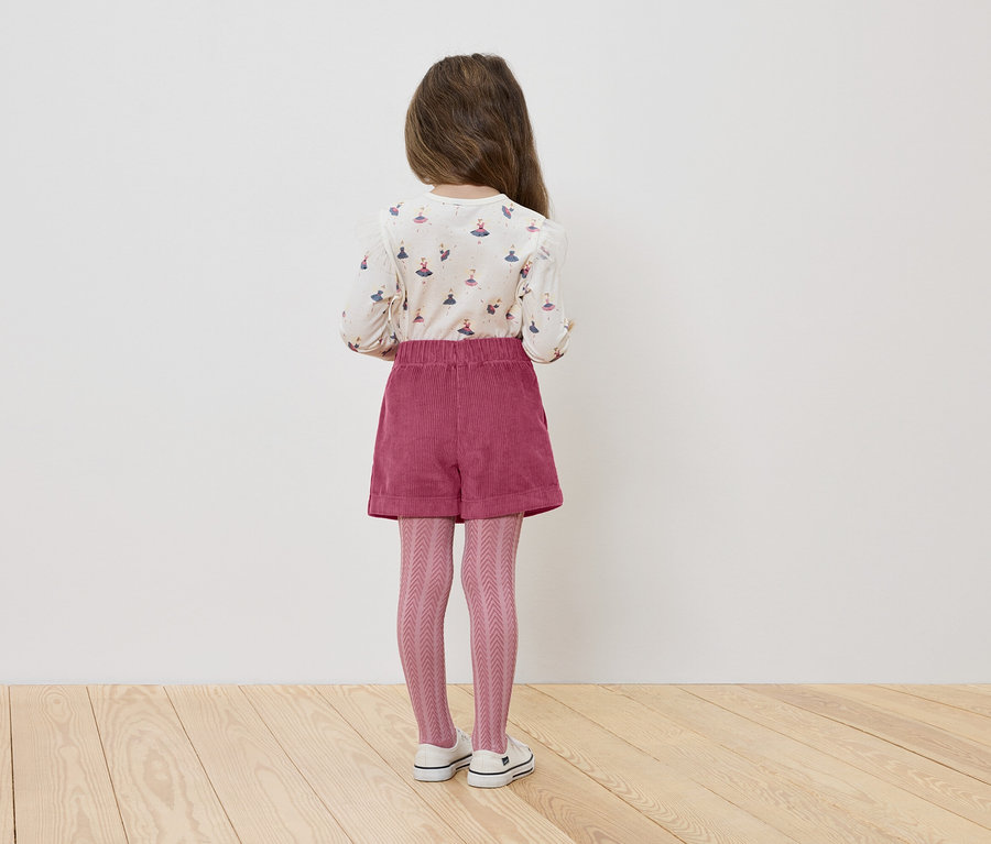 Egy hátulról látható kislány balerinás pólót, rózsaszín rövidnadrágot és kötött harisnyát visel.