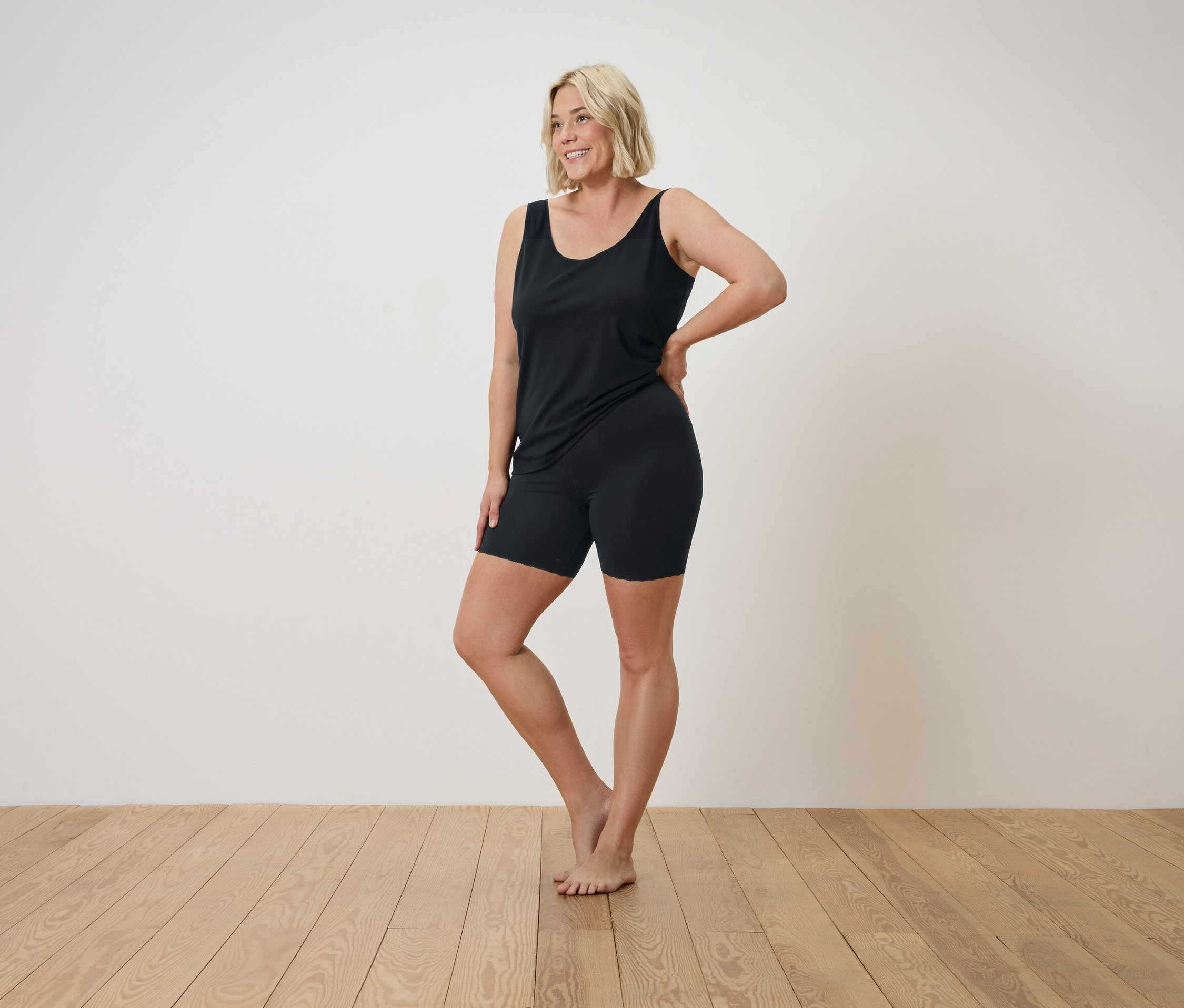 Szőke nő fekete trikóban és rövidnadrágban pózol a fapadlón.