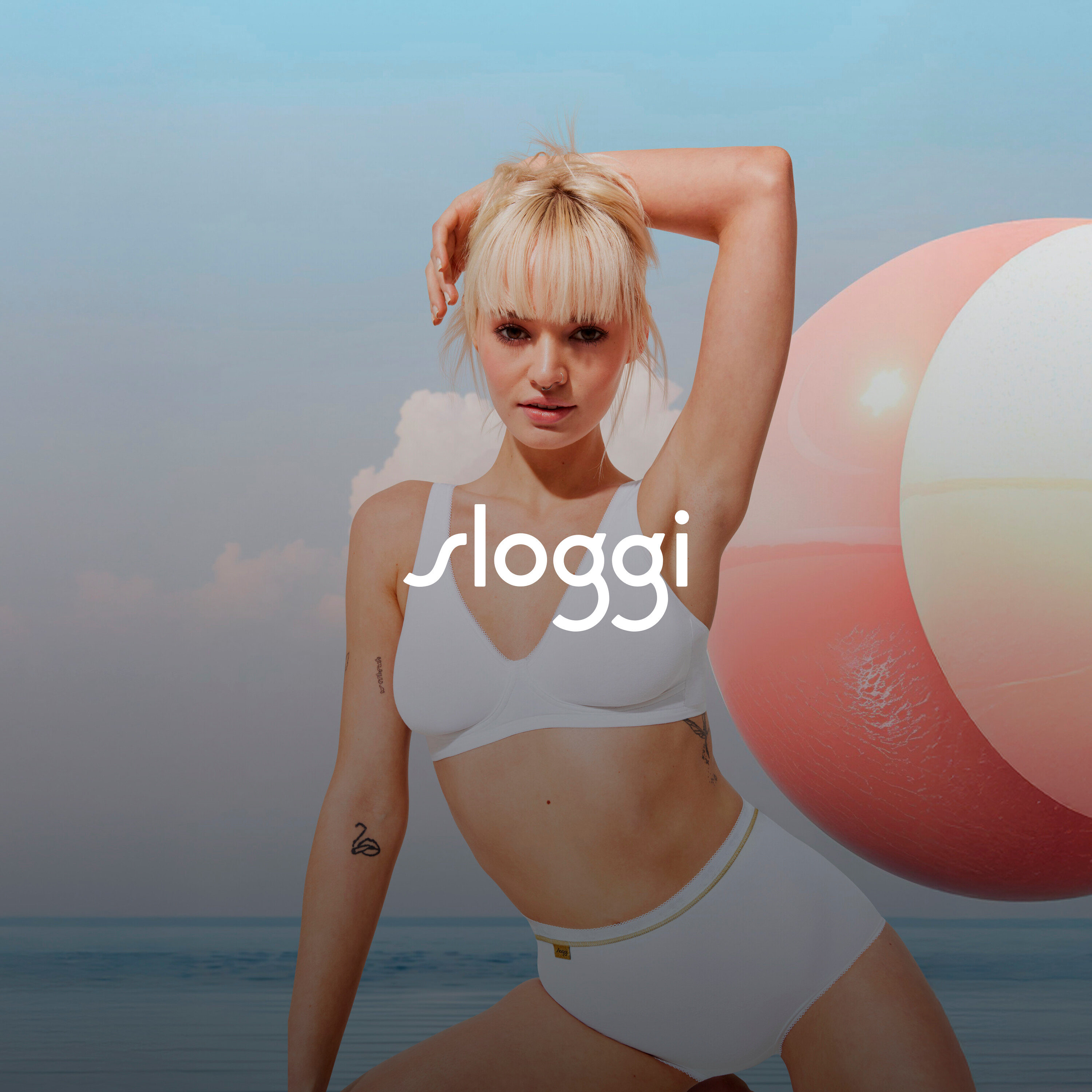 Szőke nő fehérneműben pózol felemelt kézzel egy strandlabda mellett.