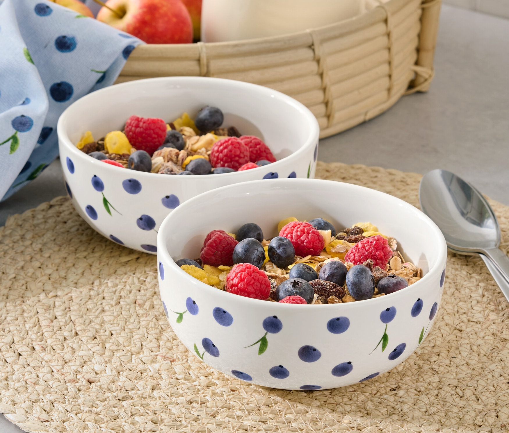 Két tál müzli málnával és áfonyával egy tányéralátéten. A háttérben egy kancsó tej és almák egy kosárban.