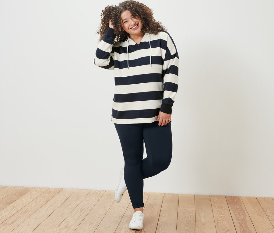 Nő pózol csíkos kapucnis szabadidőfelsőben és sötétkék leggingsben.
