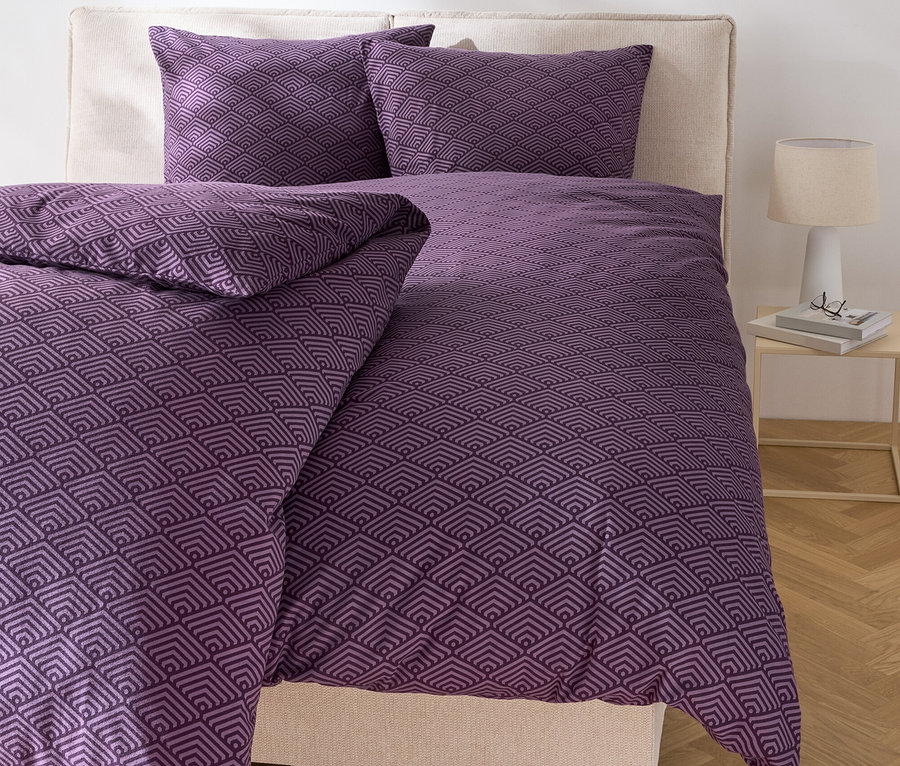 Ágy lila, geometrikus ágyneművel és két párnával.