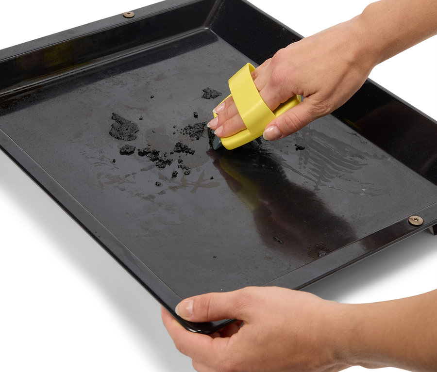 Kezek tisztítanak egy fekete sütőlemezt egy sárga tisztító eszközzel.