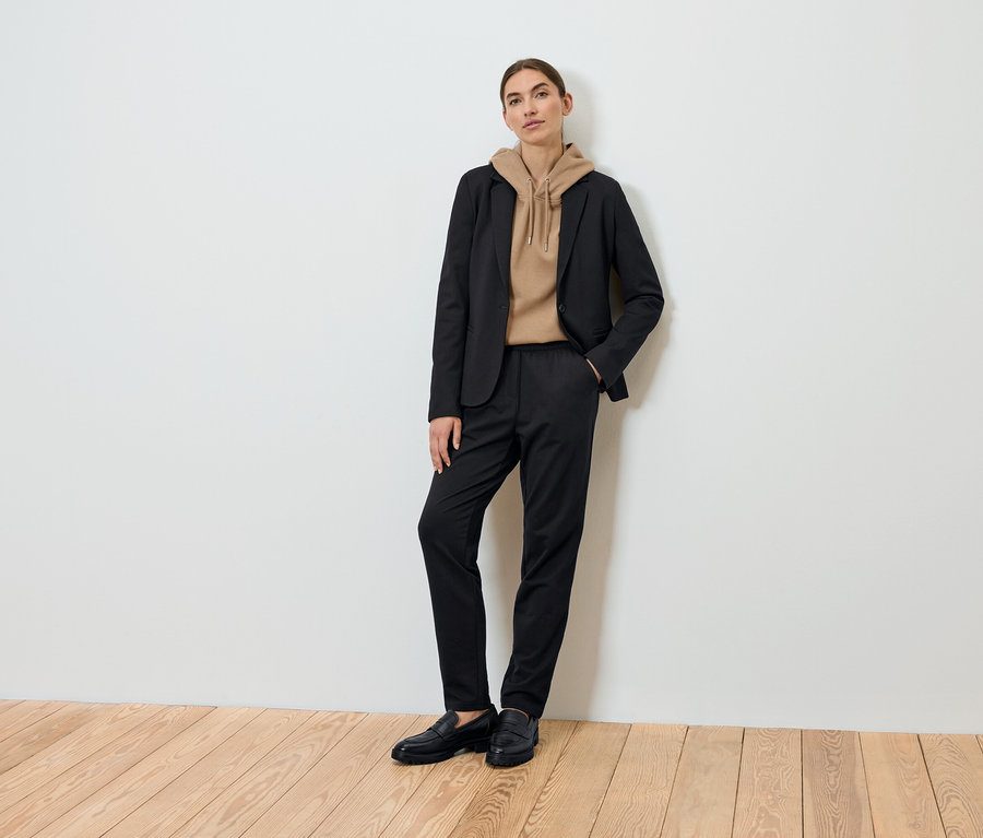 Egy nő pózol fekete blézerben, barna kapucnis pulóverben és fekete nadrágban.