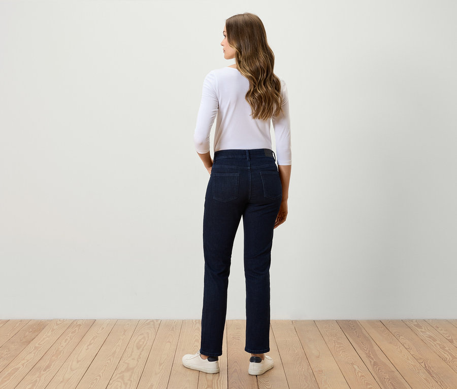 Egy nő áll háttal, sötét farmerben, fehér háromnegyedes ujjú pólóban és fehér tornacipőben.