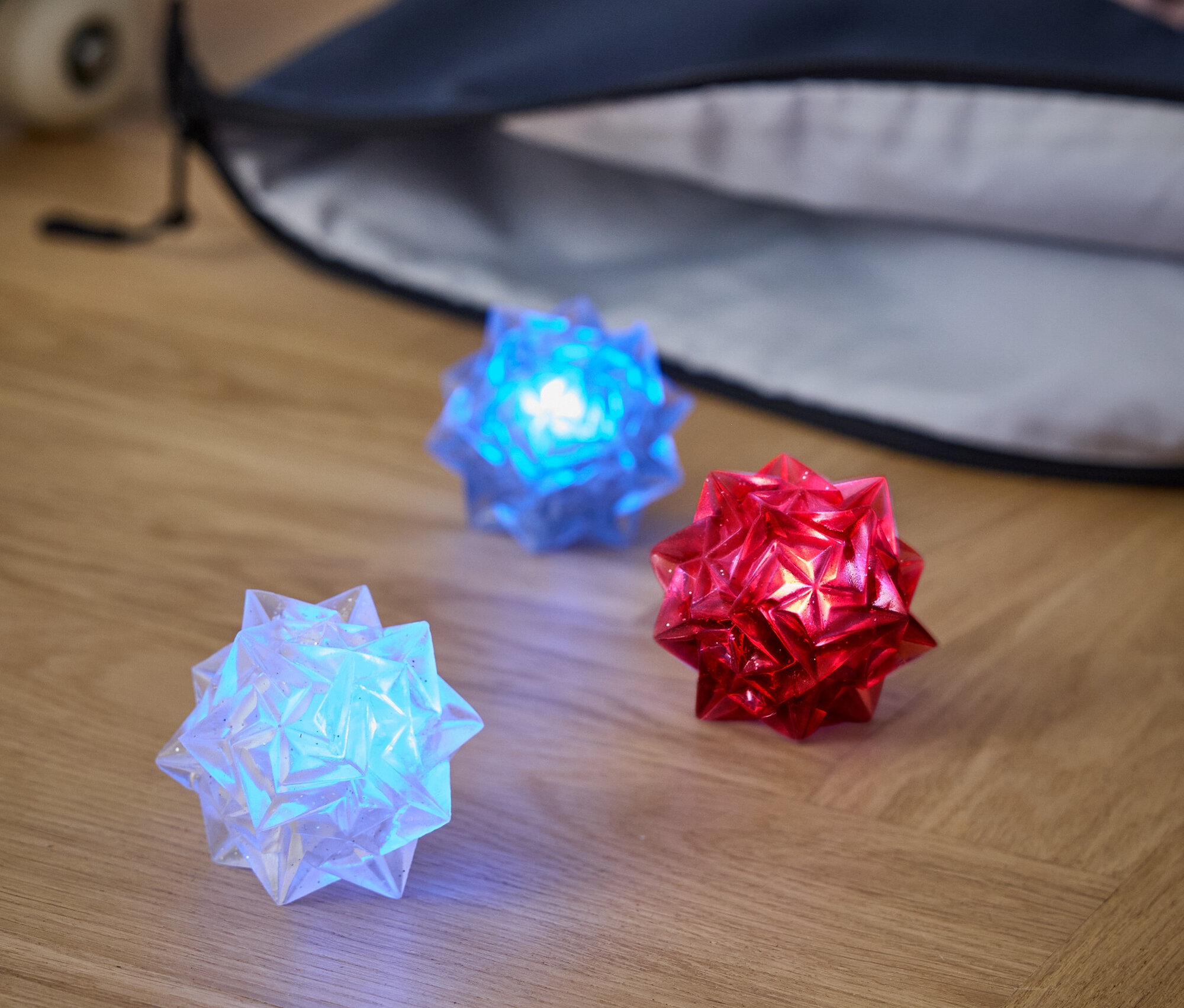 Három világító gömb fekszik egy fa padlón: egy fehér, egy kék és egy piros. Mögöttük egy hátizsák és egy gördeszka látható.