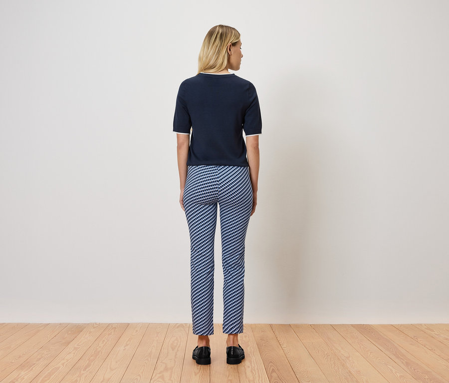 Egy szőke nő áll háttal a kamerának, sötétkék rövid ujjú felsőt és kék-fehér csíkos nadrágot visel. Fapadlón áll.