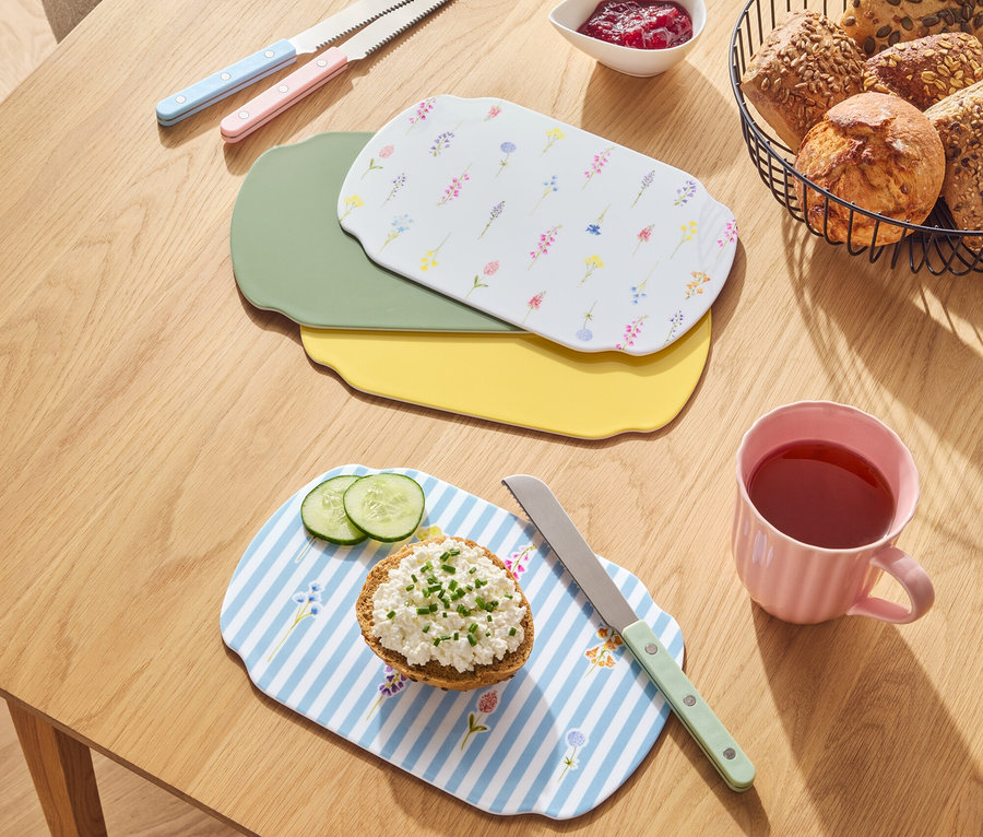 Egy fa asztalon kenyerek vannak egy kosárban, lekvár, kések és különböző színű vágódeszkák.