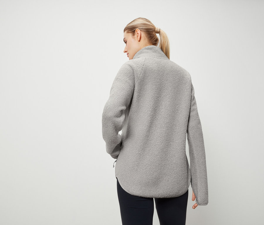 Hátulról egy szőke hajú, lófarokba kötött nő látható, aki szürke polár pulóvert és sötétkék nadrágot visel.