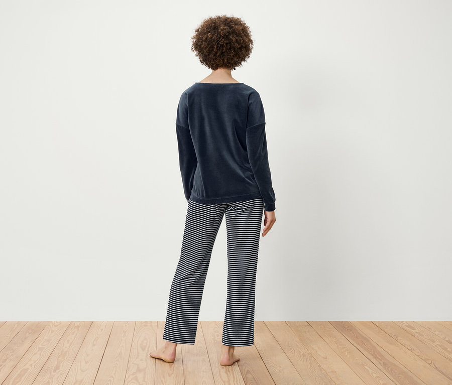 Egy göndör hajú nő háttal áll, sötétkék bársony felsőt és csíkos nadrágot visel.