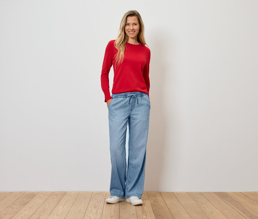 Egy nő piros pulóverben és világoskék, bő szárú nadrágban áll egy fapadlón.